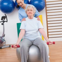 Therapist helping senior woman fit dumbbells on exercise ball in fitness studio